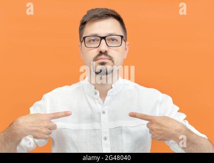 Bearded handsome man in eyeglasses pointing oneself with fingers. Shows himself, a confident calm guy points with two fingers at himself isolated on yellow color background.  Stock Photo