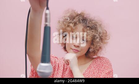 Attractive young girl holding a microphone through its wire. The mic is hanging upside down. Girl thinking. Dressed in a pink casual top. Isolated over pink background studio. Stock Photo