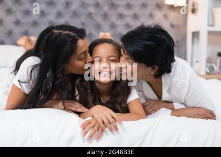 Grandmother and mother kissing smiling girl while lying on bed Stock Photo