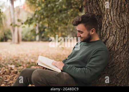 Young Caucasian man sitting next to the tree and reading a book Stock Photo