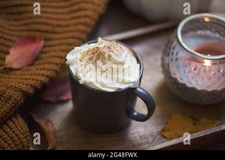 Cozy autumn cup of coffee with cream and cinnamon, blanket and candle on wooden table Stock Photo