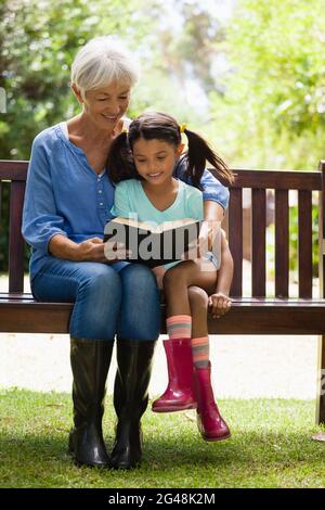 Smiling grandmother and granddaughter reading novel while sitting on wooden bench Stock Photo