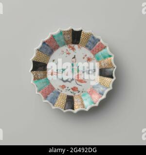 Saucer with a Chinese Lady in A Garden and Diaper Patterns on Various Colored Grounds. Dish of porcelain with ribbed wall and scalloped edge, painted on the glaze in blue, red, pink, green, yellow, black and gold. On the platter of the dish a Chinese lady (long lice) sitting on a rock in a fenced garden with flowering plants; The edge divided into narrow courses with napkin on different colored backgrounds. Family Rose. Stock Photo