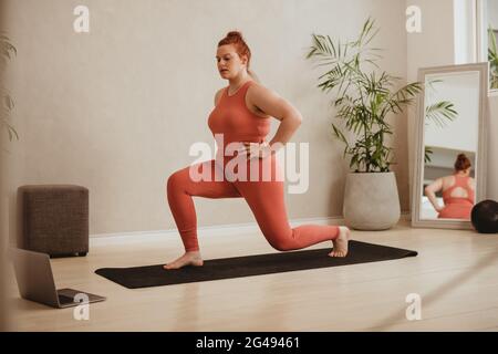 Plus size female in sportswear doing lunge workout watching online exercise class. Woman exercising over online workout class on her laptop. Stock Photo