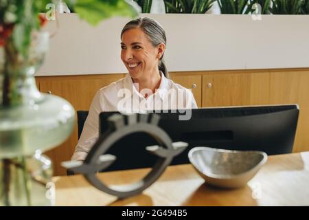 Mature woman sitting at office front desk and smiling. Happy mature office receptionist looking away and smiling. Stock Photo