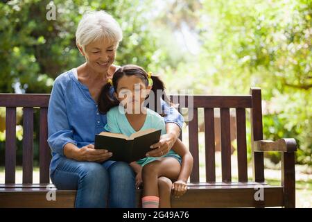 Grandmother reading novel to granddaughter sitting on wooden bench Stock Photo