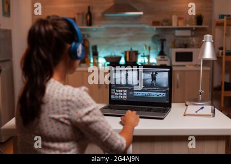 Brunette video editor works with footage on personal laptop in home ktichen during night time. Content creator in home working on montage of film using modern software for editing late at night. Stock Photo