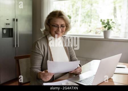 Happy older woman busy working on laptop with paperwork Stock Photo