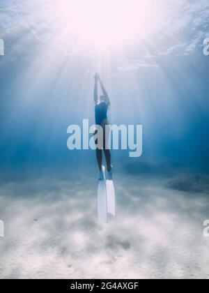 Female freediver glides with fins underwater in ocean with sunlight. Stock Photo
