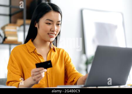 Online shopping. Positive young asian brunette woman sit at a table, uses laptop and credit card to pay for online purchases and delivery, enters details, looks at the card, smiles. Online transaction Stock Photo
