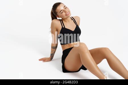 Female athlete in sports clothing, resting on floor after workout jogging, isolated on white background. Sportswoman sitting in activewear with Stock Photo