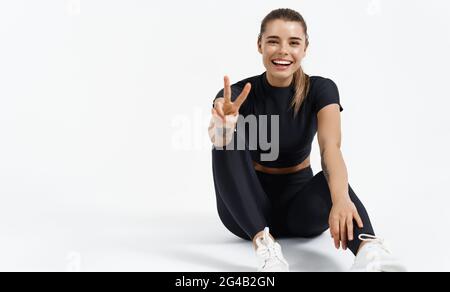 Sport and gym. Young happy athlete female sits on floor in black sportswear, show peace v-sign, smiling happy. Sporty strong woman resting after Stock Photo