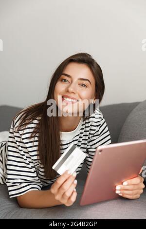 Vertical shot of smiling happy woman lying on sofa at home with tablet and credit card, online shopping, order smth in internet or paying for pucrhase Stock Photo