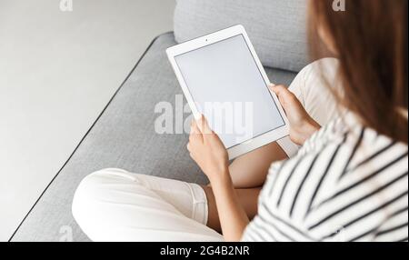 Cropped shot of woman, hands holding digital tablet with white screen, mockup promotion empty space on display, sitting with crossed legs on sofa at Stock Photo