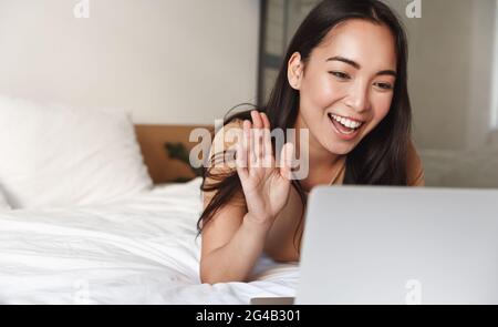 Asian girl lying in bed and video chat on laptop, waving at computer camera to say hello, greeting people online conference, communicating remotely Stock Photo
