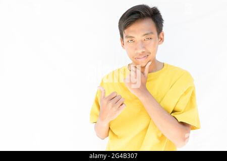 An Indonesian guy with a yellow t-shirt showing thumbs up and thinking on a white background Stock Photo