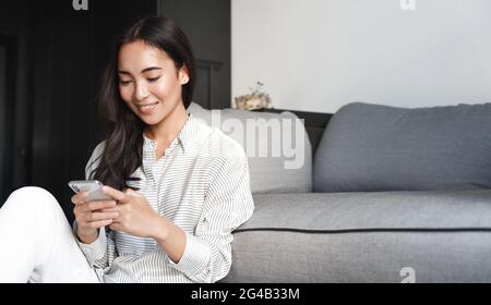 Image of smiling asian woman using smartphone at home. Girl student sitting on living room floor and reading message on mobile phone, reading screen Stock Photo