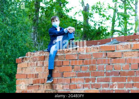 A teenage boy in a blue shirt, jeans and a medical protective mask during the coronavirus pandemic sits on a brick wall. Selective focus Stock Photo