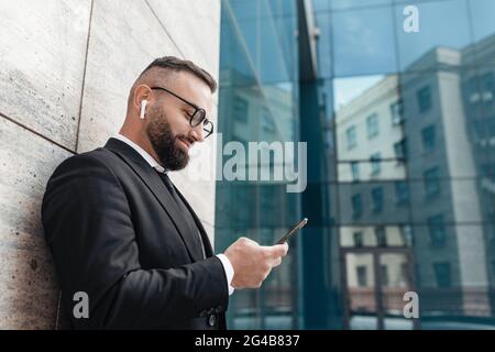 Happy mature businessman listening to music with wireless earphones and smartphone, leaning on business center wall Stock Photo