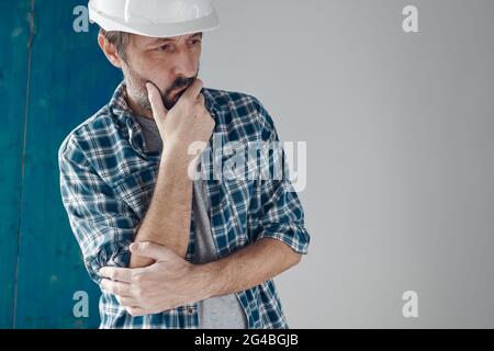 Thoughtful construction foreman in holding chin pose. Adult caucasian male with white hard hat. Stock Photo