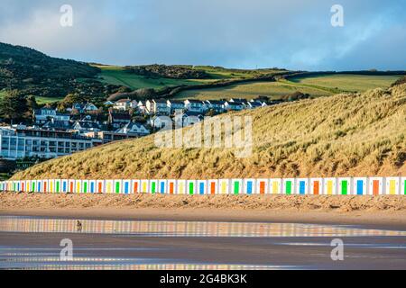 White beach huts, with coloured doors, stand in front of dunes with reflections in the wet beach sand on Woolacombe Beach, Devon Stock Photo