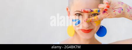 Playful portrait of a young gorgeous female artist painter covered in paint, looking and smiling at camera through her fingers. Creativity and individ Stock Photo