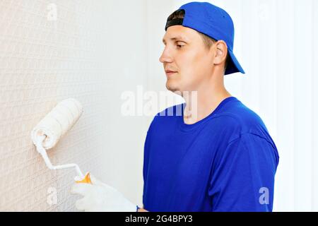 A painter in a cap and gloves paints the wall with a paint roller. Portrait of a young worker in uniform. Professional repair of premises. Mockup Stock Photo