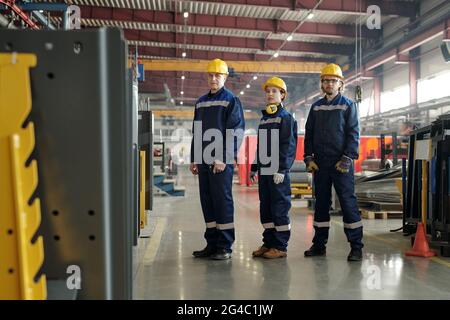 Group of confident workers of compressor plant wearing yellow hardhats and blue workwear standing in row at factory workshop Stock Photo