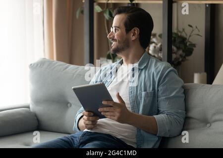 Happy dreamy young man holding digital tablet in hands. Stock Photo