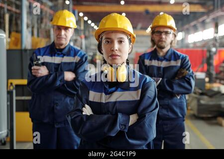 Portrait of serious young woman in hardhat and noise-cancelling headphones working with male colleagues at factory Stock Photo