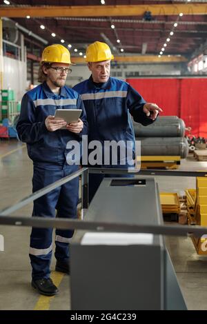 Mature foreman in hardhat showing manufacturing equipment to young specialist with tablet while explaining his tasks Stock Photo