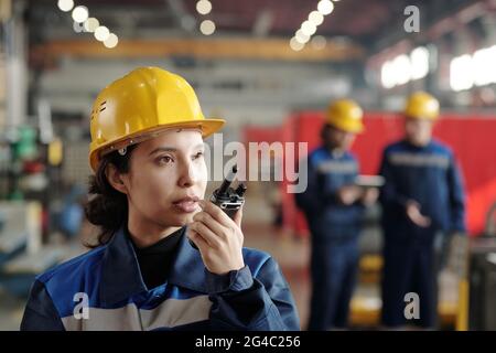 Serious young mixed race factory woman in yellow hardhat transmitting message through walkie-talkie in industry Stock Photo