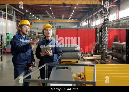 Bearded factory engineer in hardhat and safety goggles pointing at chains and explaining coworker how to use industrial crane Stock Photo