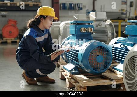 Serious young mixed race engineer in hardhat crouching at wooden pallet and using digital tablet while inspecting compressor unit Stock Photo