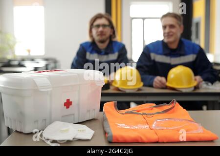Close-up of table with first aid kit for factory workers, orange vest and respiratory mask, men with hardhats in background Stock Photo