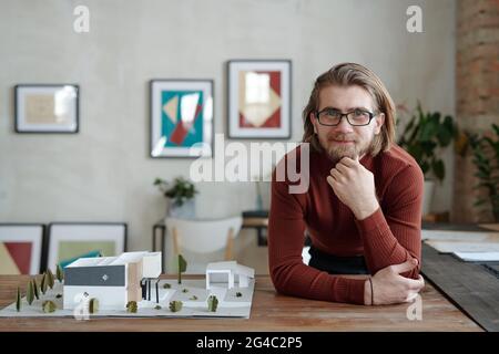 Portrait of smiling young Caucasian architect in eyeglasses leaning on table with 3D model of contemporary building in office Stock Photo
