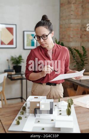 Confident young Hispanic woman in eyeglasses standing at desk with maquette of modern building and analyzing house plan Stock Photo