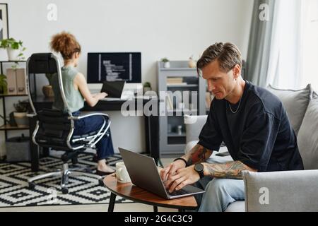 Mature businessman sitting on sofa and working on laptop with his colleague in the background Stock Photo