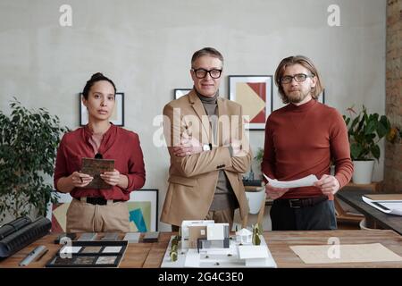 Portrait of confident multi-ethnic architects in smart casual outfits standing at table with samples and maquette while working on new design Stock Photo