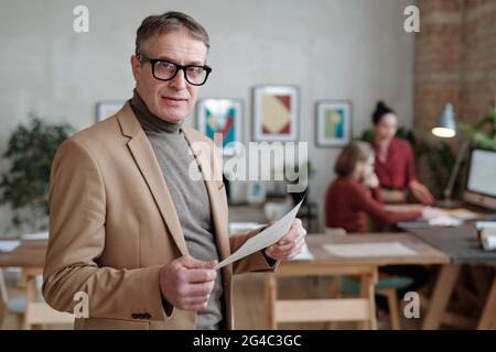 Portrait of content confident mature businessman in eyeglasses holding paper and standing against working colleagues in background Stock Photo
