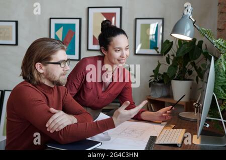 Positive young interracial architects in burgundy outfits looking at computer monitor and discussing online sketch in office Stock Photo