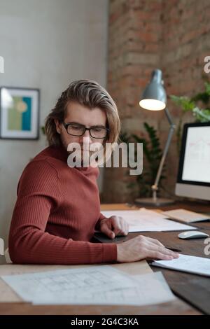 Focused young Caucasian bearded architect in glasses sitting at table and looking at sketch while examining it in office Stock Photo