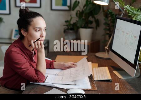 Serious young mixed race woman with hair bun sitting at desk and holding house plan while analyzing design sketch on computer Stock Photo