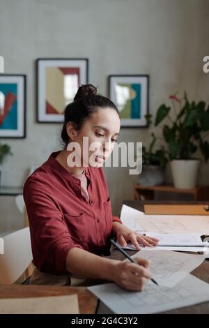 Concentrated young Hispanic woman in burgundy shirt sitting at desk and drawing sketch while developing building design Stock Photo
