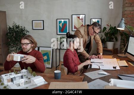 Young architect discussing blueprint with chief while bearded designer working with 3D model of cottage house in office Stock Photo