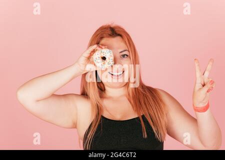 Cheerful young redhead plus size woman holding sweet donut near her eye looking through and showing peace gesture while standing on pink background. Stock Photo