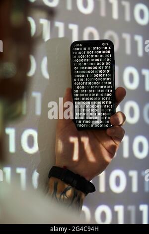 Rear view of hacker holding mobile phone and examining computer code on it Stock Photo