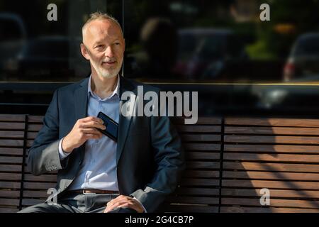 The man in a gray suit with a smartphone sitting on a bench. Stock Photo