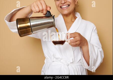 Smiling mature woman with toothy smile in white bathrobe pouring coffee drink from a geyser coffee maker into a glass cup. Isolated portrait over beig Stock Photo