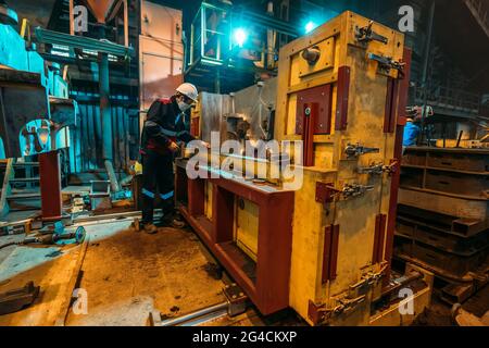 Unrecognizable worker in uniform, helmet and protective face mask works at machine in workshop of metallurgical plant. Stock Photo